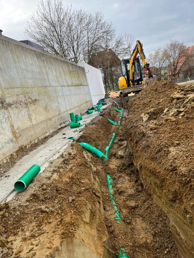 Graben mit grünem Rohrleitungen und Baggerarbeiten im Hintergrund.